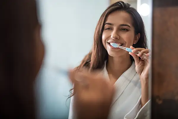 Young pregnant woman in a white robe smiling and brushing her teeth while looking into a bathroom mirror, practicing good dental hygiene.